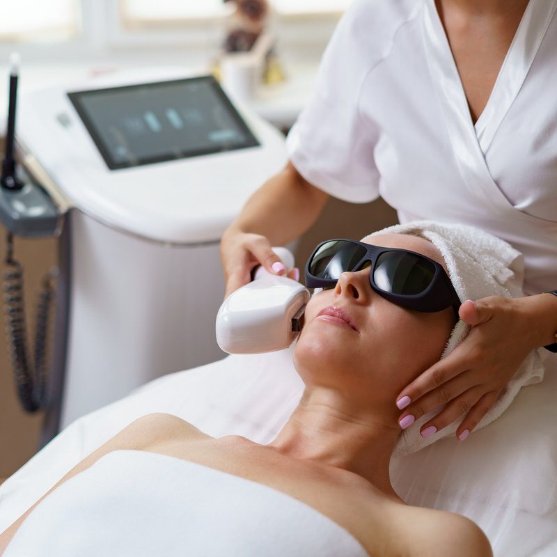 View of doctor cosmetologist doing anti aging procedure in cosmetology office. Satisfied woman in disposable hat lying on couch and relaxing. Working with Apparatus.